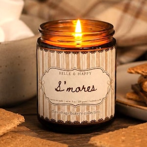 May include: A lit candle in an amber glass jar with a label reading Belle & Happy S'mores, the cottage bakery collection, and net wt. 8 oz. The label features a brown and white striped pattern with a decorative border and scalloped edges.