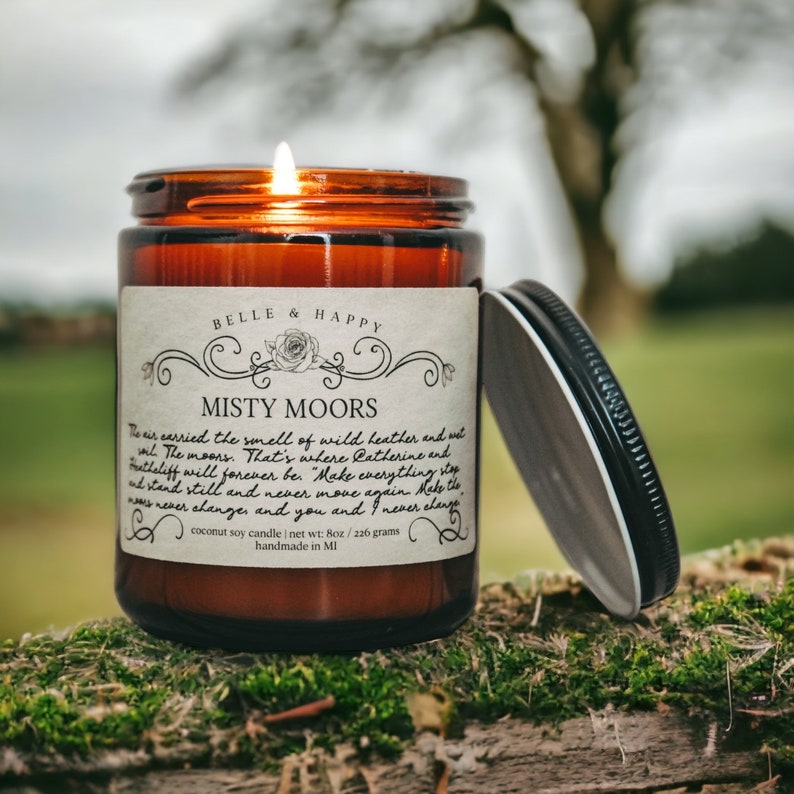 May include: A brown glass candle jar with a white label that reads "Belle & Happy Misty Moors". The label has a decorative floral design and text that reads "The air carried the smell of wild heather and wild. The moors. That's where Catherine and Heathcliff will forever be. "Make everything stop and stand still and never, never move again. Make the waves never change, and you and I never change." The candle is a coconut soy candle with a net weight of 8 oz or 226 grams. It is handmade in MI.