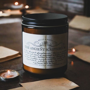 May include: A brown glass candle jar with a black lid and a white label that reads "Belle & Happy" and "The Ancient Scriptorium". The candle is surrounded by old books and a warm glow.