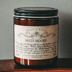 May include: A brown glass jar candle with a white label that reads "BELLE & HAPPY" and "MISTY MOORS". The label has a handwritten description of the scent and the text "coconut soy candle | net wt: 8oz / 226 gram | handmade in MI".