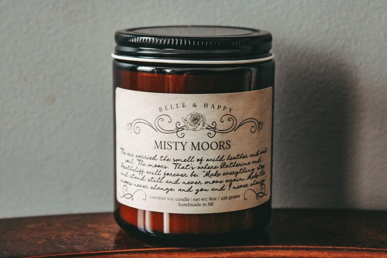 May include: A brown glass jar candle with a white label that reads "BELLE & HAPPY" and "MISTY MOORS". The label has a handwritten description of the scent and the text "coconut soy candle | net wt: 8oz / 226 gram | handmade in MI".