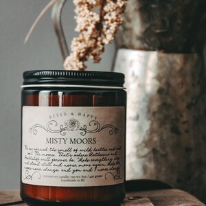 May include: A brown glass candle jar with a black lid and a white label that reads "BELLE & HAPPY" and "MISTY MOORS". The label has a floral design and text that reads "This carries the smell of wild heather and mist. The moors. That's where Catherine and Mettelliff will forever be. "Make everything stand still and never move again. Help the moors never change and you and I never change." The candle is a coconut soy candle and weighs 7 oz / 200 grams. It is handmade in MI.