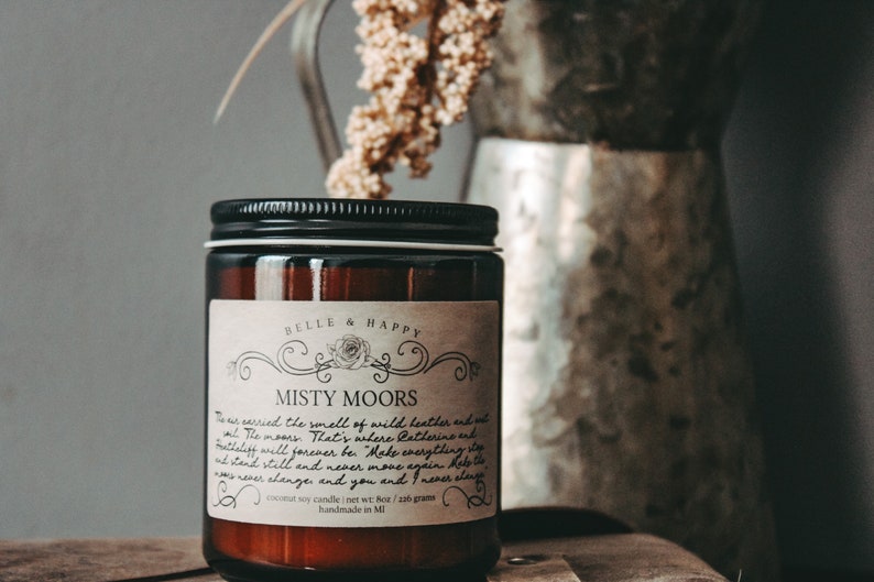 May include: A brown glass candle jar with a black lid and a white label that reads "BELLE & HAPPY" and "MISTY MOORS". The label has a floral design and text that reads "This carries the smell of wild heather and mist. The moors. That's where Catherine and Mettelliff will forever be. "Make everything stand still and never move again. Help the moors never change and you and I never change." The candle is a coconut soy candle and weighs 7 oz / 200 grams. It is handmade in MI.