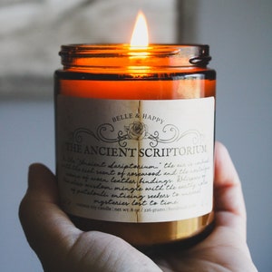 May include: A brown glass candle jar with a white label that reads "Belle & Happy" and "The Ancient Scriptorium." The label has a decorative border and a small image of a rose. The candle is lit and the flame is visible.