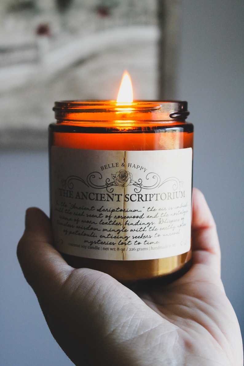 May include: A brown glass candle jar with a white label that reads "Belle & Happy" and "The Ancient Scriptorium." The label has a decorative border and a small image of a rose. The candle is lit and the flame is visible.