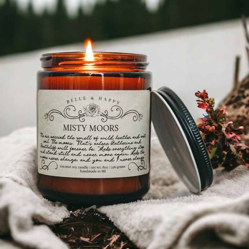 May include: A brown glass candle jar with a white label that reads "BELLE & HAPPY" and "MISTY MOORS". The label has a decorative floral design and the text "coconut soy candle net wt: 8oz / 226 grams handmade in MI".