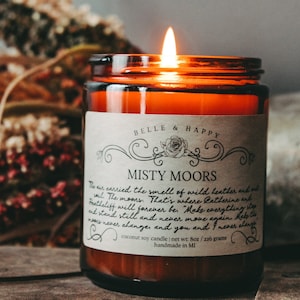 May include: A brown glass candle jar with a white label that reads "Belle & Happy Misty Moors". The label has a floral design and text that reads "The air carried the smell of wild heather and salt. The moors... That's where Catherine and Heathcliff will forever be. Make everything stand still and never move again. Help the moors never change and you and I never alone. Coconut soy candle | net wt: 8oz / 216 grams handmade in MI"