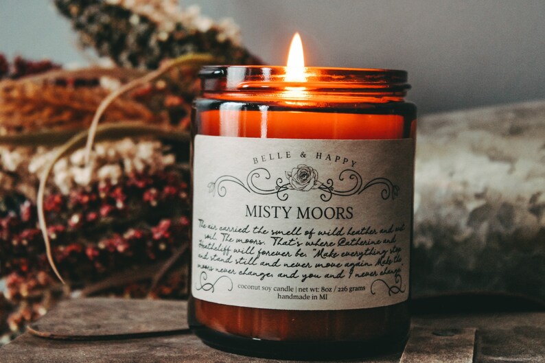 May include: A brown glass candle jar with a white label that reads "Belle & Happy Misty Moors". The label has a floral design and text that reads "The air carried the smell of wild heather and salt. The moors... That's where Catherine and Heathcliff will forever be. Make everything stand still and never move again. Help the moors never change and you and I never alone. Coconut soy candle | net wt: 8oz / 216 grams handmade in MI"