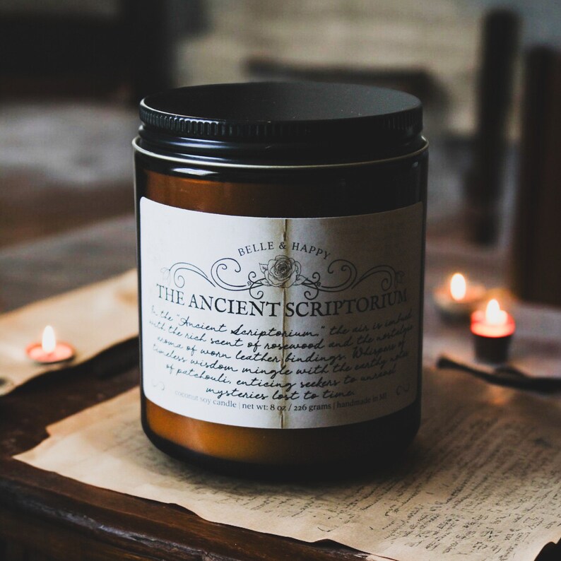 May include: A brown glass candle jar with a black lid and a white label that reads "BELLE & HAPPY THE ANCIENT SCRIPTORIUM". The label has a decorative design with a rose and script text. The candle is surrounded by old paper and lit tea lights.