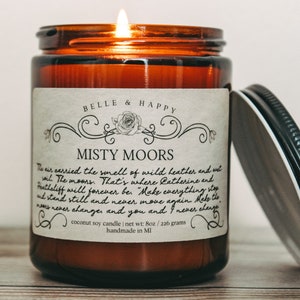 May include: A brown glass candle jar with a white label that reads "BELLE & HAPPY" and "MISTY MOORS". The label has a quote about the moors and a rose design. The candle is lit and the lid is off. The candle is on a wooden surface.