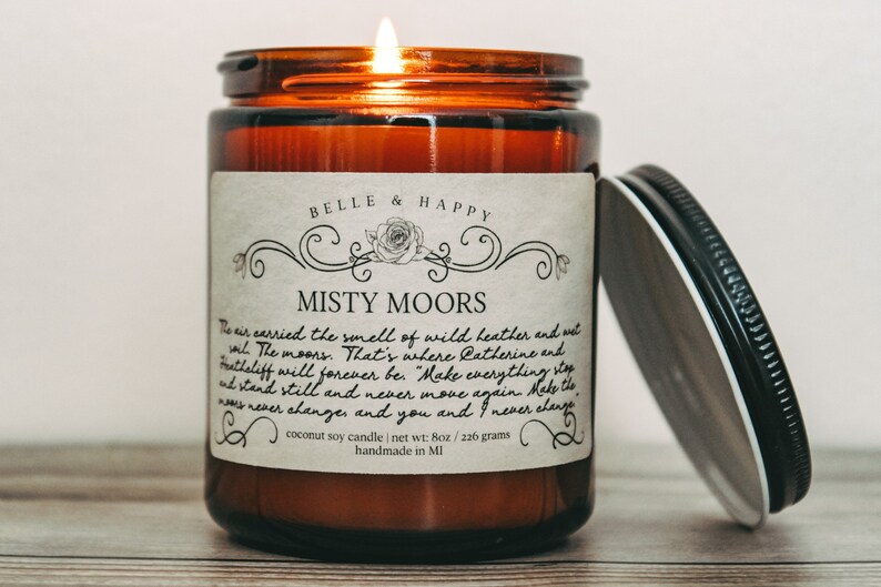 May include: A brown glass candle jar with a white label that reads "BELLE & HAPPY" and "MISTY MOORS". The label has a quote about the moors and a rose design. The candle is lit and the lid is off. The candle is on a wooden surface.