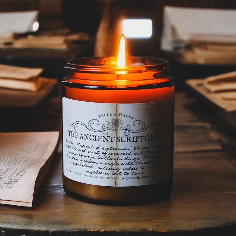 May include: A brown glass candle jar with a white label that reads "Belle & Happy The Ancient Scriptorium". The candle is lit and the flame is casting a warm glow. The candle is on a wooden surface with books in the background.