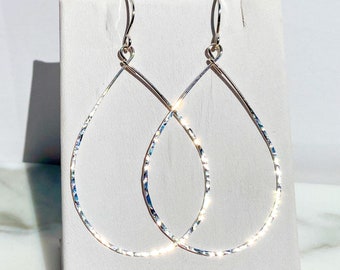 May include: A pair of silver-toned teardrop-shaped earrings featuring a hammered, reflective surface texture. The earrings are designed with thin, open wire frames and include curved ear wires for attachment, creating a light-catching effect.