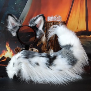 以下が含まれることがあります： 動物の耳と尻尾を模した、ヘッドバンドと尻尾のコスチュームセット。耳と尻尾は主に白で、グレーと黒の模様が入っています。ヘッドバンドは黒で、尻尾はふわふわです。