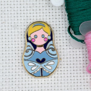 May include: A gold-colored pin with a blue and white design of a princess in a dress. The princess has blonde hair, pink cheeks, and blue eyes. The pin is shaped like a Russian nesting doll.