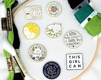May include: A collection of enamel pins with various inspirational quotes and designs. The pins are displayed on a white fabric background with green and black embroidery floss.