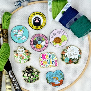 May include: A collection of nine enamel pins featuring various designs, including a cat, a turtle, a unicorn, and a dog. The pins are arranged on a white canvas with green and blue embroidery floss.