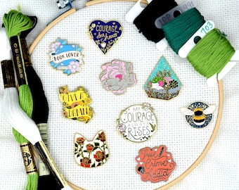 May include: A collection of enamel pins featuring various designs, including a book lover pin, a courage pin, a give a damn pin, a my courage always rises pin, a true crime addict pin, a bee pin, a terrarium pin, a cat pin, and a flower pin. The pins are displayed on a white canvas with green and black embroidery floss.