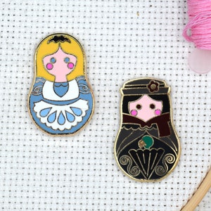 May include: Two enamel pins featuring a traditional Russian nesting doll design. The first pin is yellow with a blue and white dress and the second pin is black with a red and green dress.