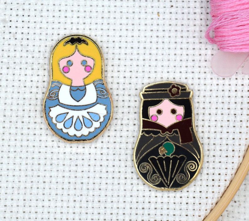May include: Two enamel pins featuring a traditional Russian nesting doll design. The first pin is yellow with a blue and white dress and the second pin is black with a red and green dress.