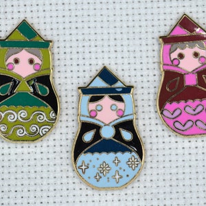 May include: Three enamel pin badges featuring cartoon women in traditional Russian nesting doll style. The dolls are wearing green, blue, and red dresses with gold trim. The dolls have pink cheeks and black eyes.