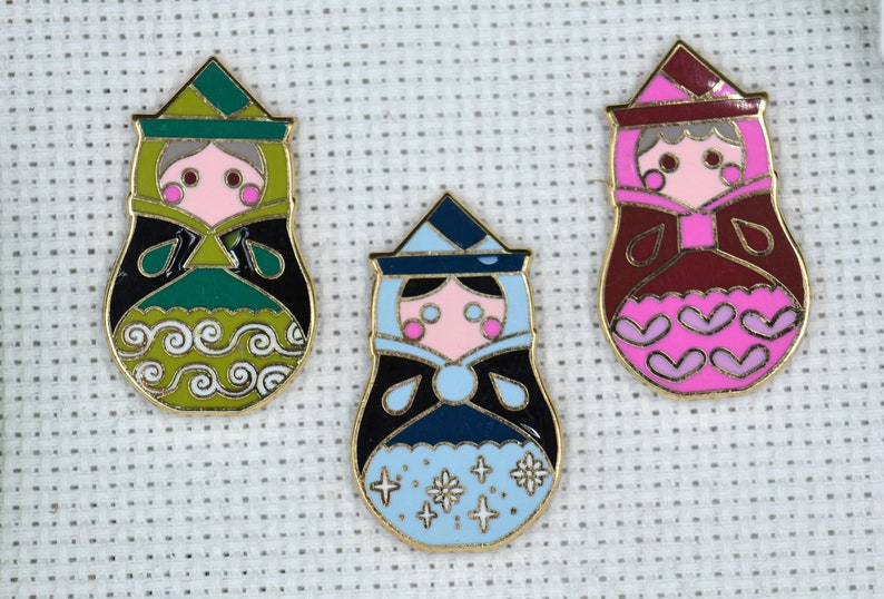 May include: Three enamel pin badges featuring cartoon women in traditional Russian nesting doll style. The dolls are wearing green, blue, and red dresses with gold trim. The dolls have pink cheeks and black eyes.