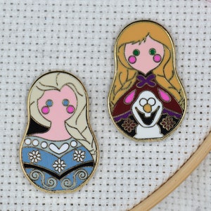 May include: Two gold-colored enamel pins featuring characters from the movie Frozen. The first pin is a round, blue and white pin with a depiction of Elsa. The second pin is a round, red and gold pin with a depiction of Anna.