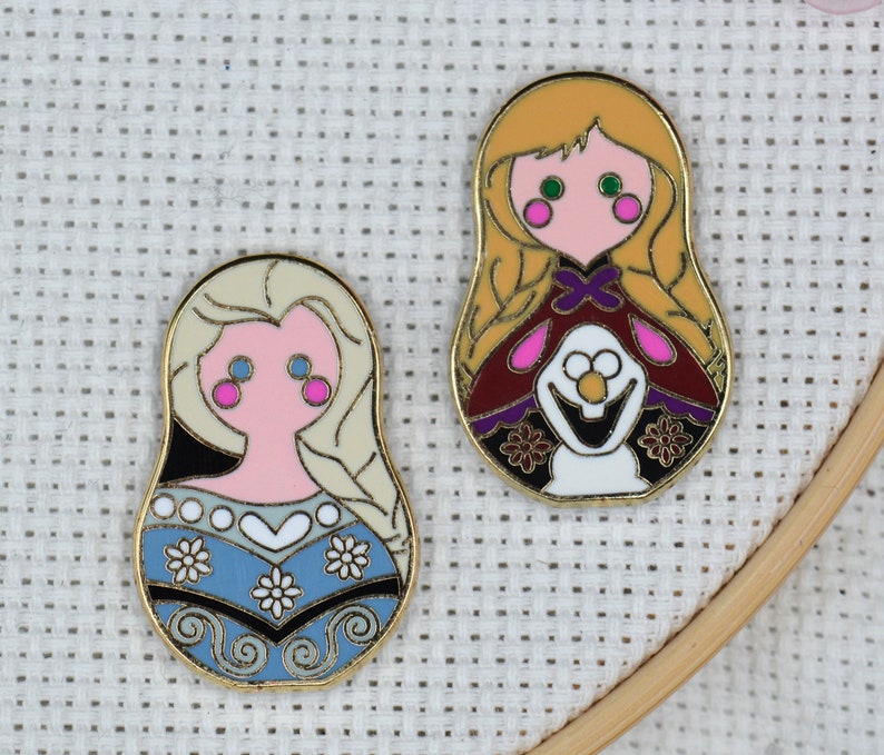 May include: Two gold-colored enamel pins featuring characters from the movie Frozen. The first pin is a round, blue and white pin with a depiction of Elsa. The second pin is a round, red and gold pin with a depiction of Anna.