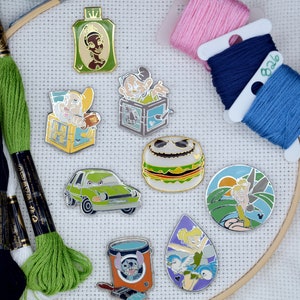 May include: A collection of enamel pins featuring various cartoon characters and designs, including a green car, a hamburger with a skull face, and a paint bucket with a character. Embroidery floss in green, pink, and blue is also visible.