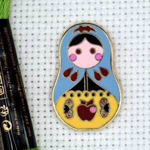 May include: A blue and yellow enamel pin featuring a stylized Russian nesting doll with a red apple and butterfly designs. The pin is on a white background.