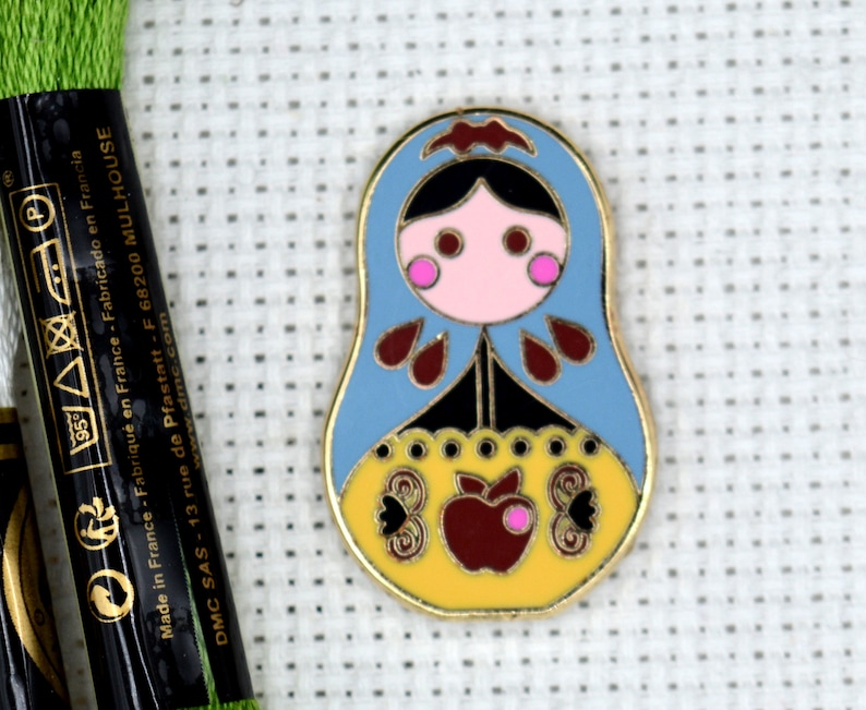 May include: A blue and yellow enamel pin featuring a stylized Russian nesting doll with a red apple and butterfly designs. The pin is on a white background.