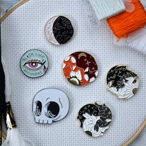 May include: Five enamel pins with different designs. The pins are on a white fabric background. The pins feature a skull, a black eye with the text "Black Like My Heart", a yin yang design with a fox and a moon, and two yin yang designs with bunnies and moons.