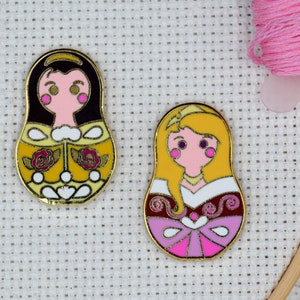 May include: Two gold-colored enamel pins featuring Disney princesses Belle and Aurora in a Russian nesting doll style. Belle is wearing a yellow dress with roses and Aurora is wearing a pink dress with a white bodice.