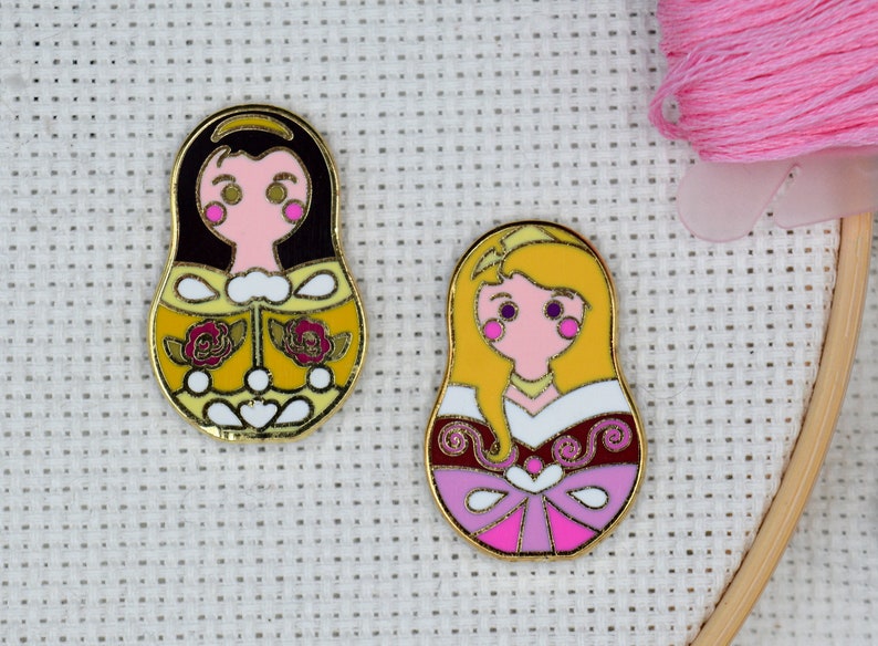 May include: Two gold-colored enamel pins featuring Disney princesses Belle and Aurora in a Russian nesting doll style. Belle is wearing a yellow dress with roses and Aurora is wearing a pink dress with a white bodice.