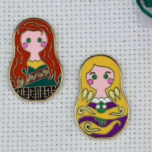 May include: Two gold-colored metal pins shaped like Russian nesting dolls. The doll on the left has red hair, green eyes, and a green, gold, and black dress. The doll on the right has yellow hair, green eyes, and a purple, gold, and green dress.
