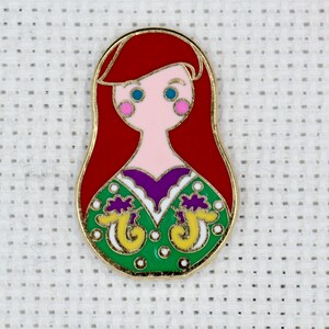 May include: A red, white, green, yellow, and purple enamel pin featuring a cartoon image of Ariel from The Little Mermaid. The pin is shaped like a Russian nesting doll.