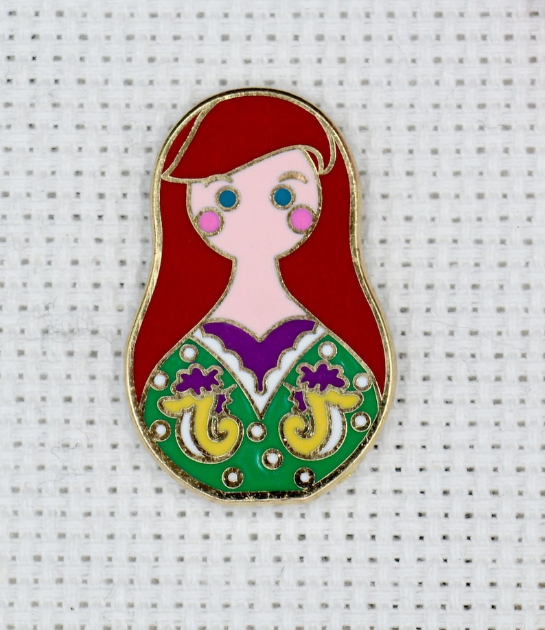 May include: A red, white, green, yellow, and purple enamel pin featuring a cartoon image of Ariel from The Little Mermaid. The pin is shaped like a Russian nesting doll.