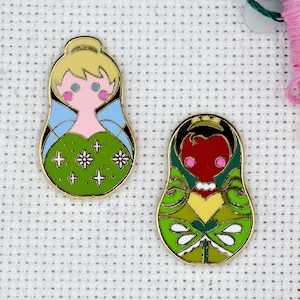 May include: Two gold-colored enamel pins featuring a green and yellow design. The first pin depicts a fairy with blonde hair, blue wings, and a pink dress. The second pin depicts a princess with black hair, a yellow crown, and a red dress.