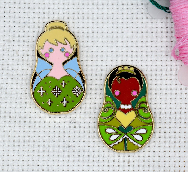 May include: Two gold-colored enamel pins featuring a green and yellow design. The first pin depicts a fairy with blonde hair, blue wings, and a pink dress. The second pin depicts a princess with black hair, a yellow crown, and a red dress.