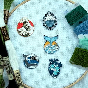 May include: Five enamel pins with ocean themes. The pins are on a white canvas background with green, blue, and white embroidery floss. The pins feature a wave, a whale, a bottle in the ocean, a whale tail, and a heart.