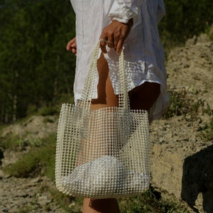 May include: A white mesh tote bag with a white fabric lining. The bag is being held by a person wearing a white shirt.