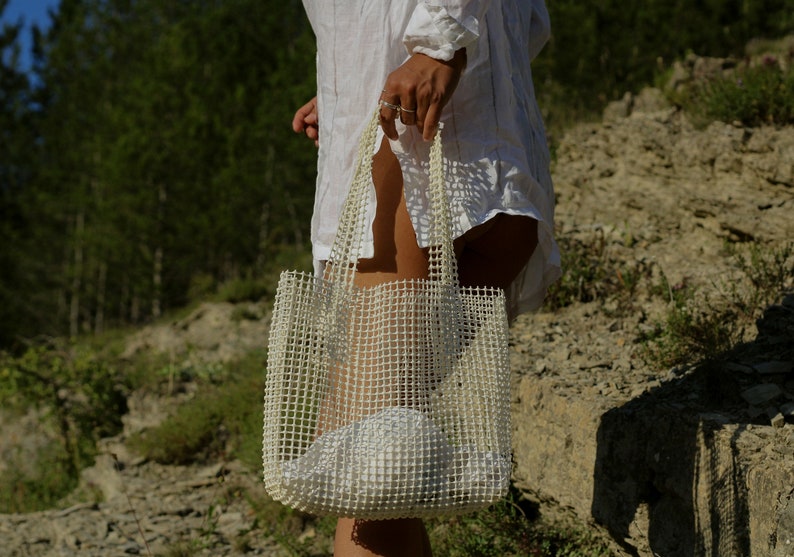 May include: A white mesh tote bag with a white fabric lining. The bag is being held by a person wearing a white shirt.