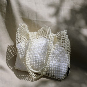 May include: A white mesh tote bag with a white fabric lining. The bag is folded and sitting on a beige fabric surface.