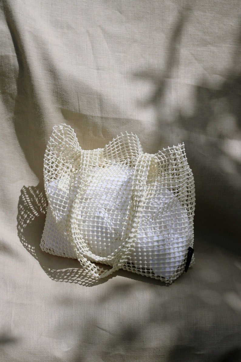 May include: A white mesh tote bag with a white fabric lining. The bag is folded and sitting on a beige fabric surface.