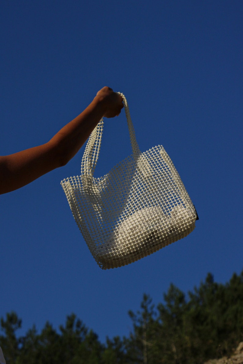 May include: A white mesh tote bag with a white fabric lining. The bag is being held by a hand against a blue sky.