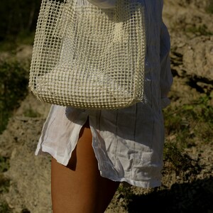 May include: A white mesh tote bag with a white shirt. The bag is being held by a person.