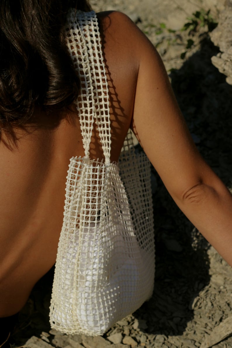 May include: A white mesh tote bag with a wide, flat base and two straps. The bag is made of a woven material with a grid pattern.