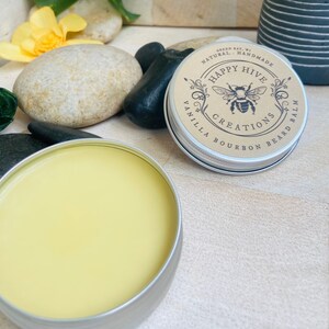 May include: A round tin container with a yellow balm inside. The lid of the tin has a brown label with the text "Happy Hive Creations Vanilla Bourbon Beard Balm" and a black and white illustration of a bee.