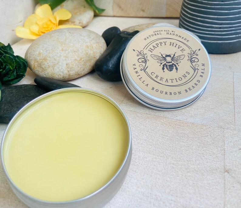 May include: A round tin container with a yellow balm inside. The lid of the tin has a brown label with the text "Happy Hive Creations Vanilla Bourbon Beard Balm" and a black and white illustration of a bee.