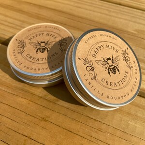 May include: Two small metal tins with brown paper labels. The labels feature a bee illustration and the text "Happy Hive Creations" with the words "Vanilla Bourbon Beard Balm" printed on the bottom of the label.